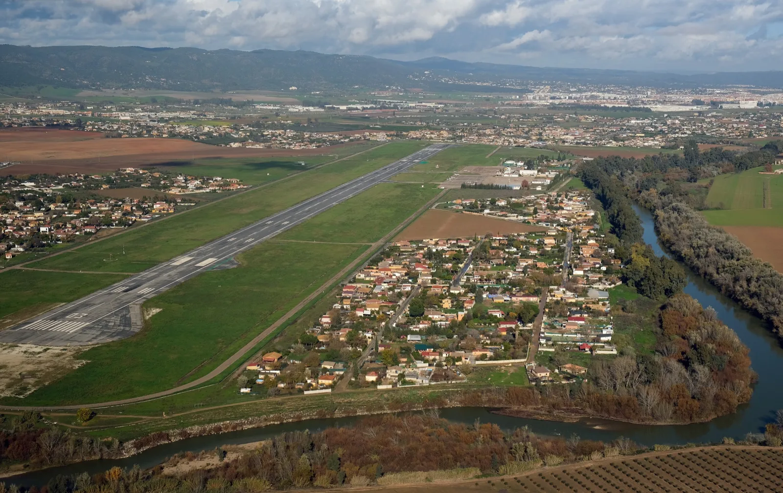 Vista aérea del aeropuerto de Córdoba entre campos verdes, con el Guadalquivir describiendo una curva y la ciudad y la sierra al fondo.