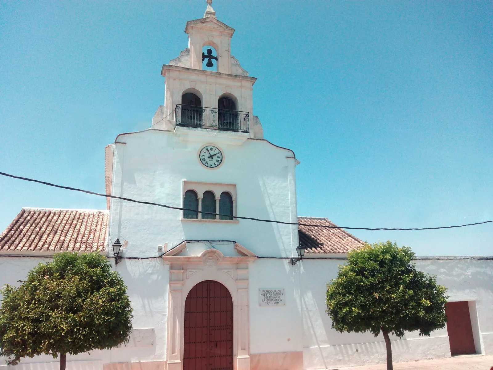 Fachada encalada de la iglesia de Nuestra Señora del Rosario de La Guijarrosa, con espadaña y reloj, puerta de madera y dos naranjos a los lados, bajo un cielo azul.