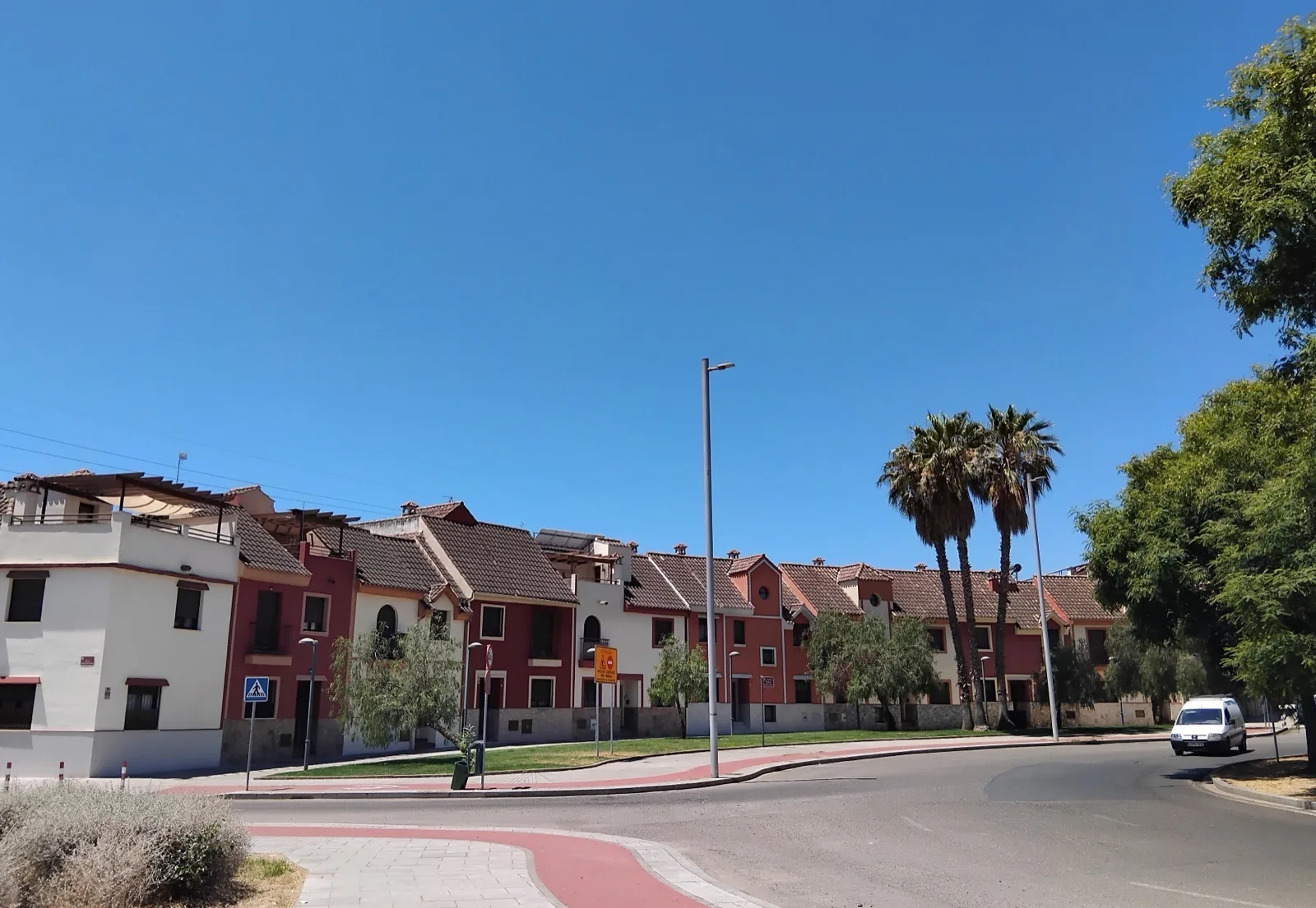 Hilera de casas adosadas de dos plantas pintadas de blanco y granate, con tejados de teja, junto a una calle con carril bici rojo, farolas y dos palmeras bajo un cielo azul.