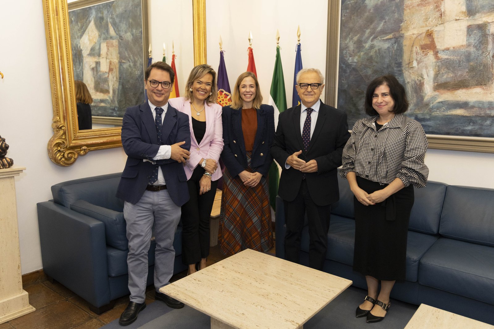 Cinco personas de pie, sonriendo, en una sala con sofás azules, banderas y cuadros.