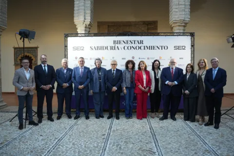 Doce personas trajeadas posan en fila ante un cartel que dice Bienestar, Sabiduría y Conocimiento en un patio con arcos.