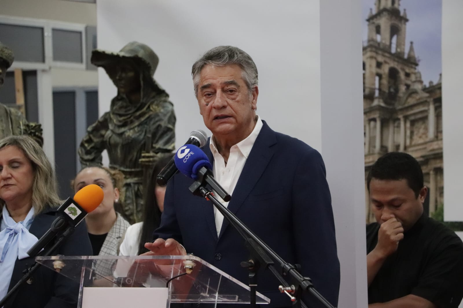 Un hombre mayor con traje azul habla ante micrófonos en un atril, con una escultura y un cartel de un edificio al fondo.
