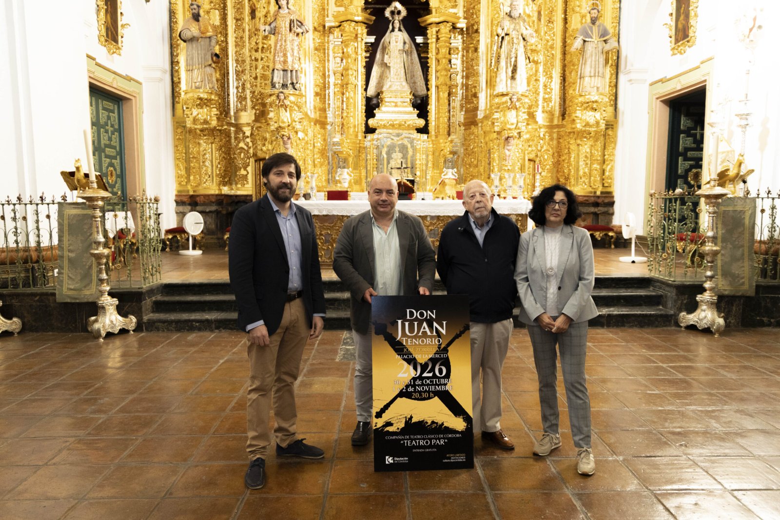 Cuatro personas de pie sostienen un cartel de Don Juan Tenorio ante un retablo dorado en el interior de una iglesia.
