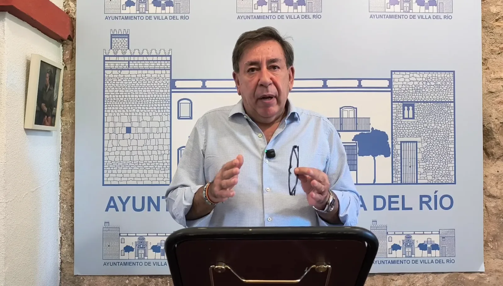 Un hombre de camisa clara gesticula tras un atril ante un cartel con dibujos y el texto Ayuntamiento de Villa del Río.