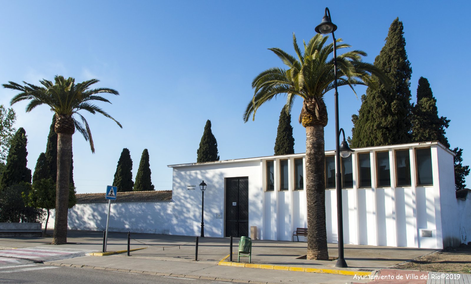 Edificio blanco de una sola planta con puerta enrejada y ventanas alargadas, junto a dos palmeras y cipreses, con una acera y una farola delante.