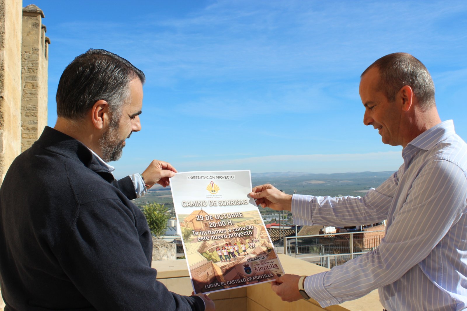 Dos hombres sostienen y miran un cartel sobre un proyecto, con un paisaje de campo al fondo.