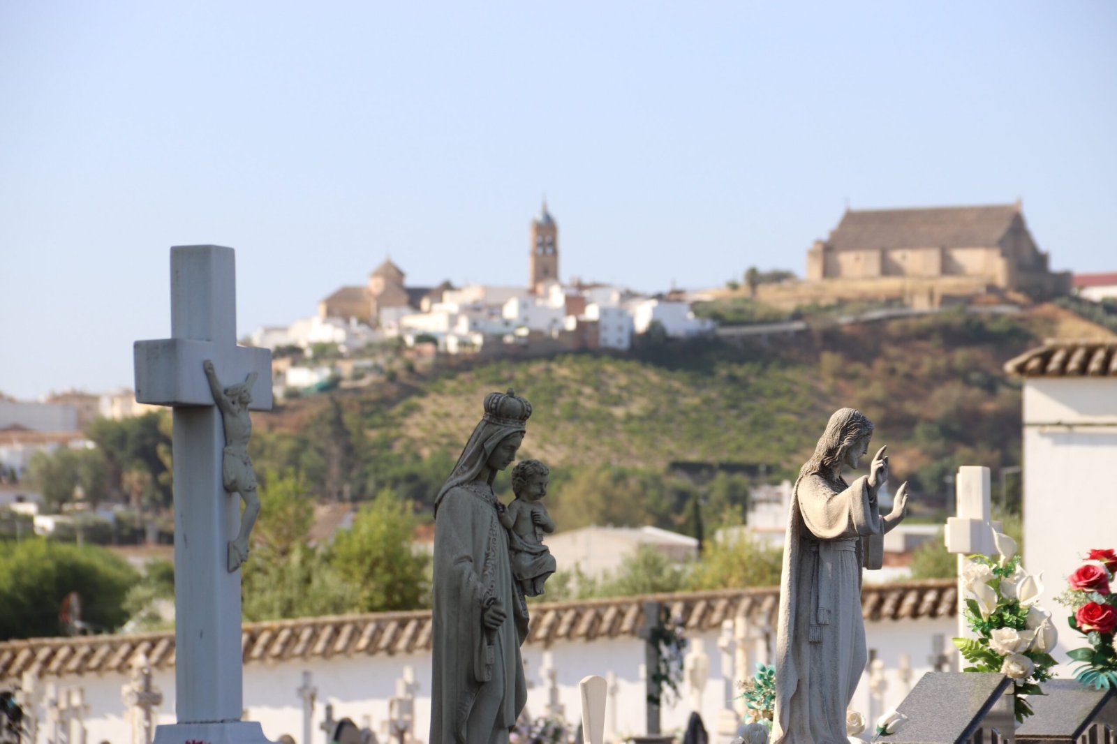 Estatuas religiosas y una cruz en un cementerio, con un pueblo blanco y una iglesia en lo alto de un cerro al fondo.
