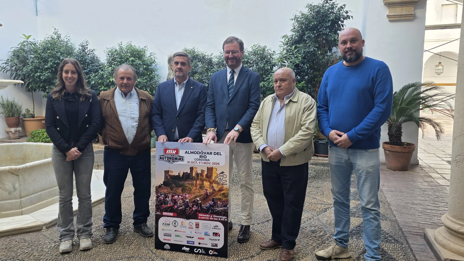 Seis personas de pie en un patio con plantas sostienen un cartel de un campeonato de motos en Almódovar del Río.