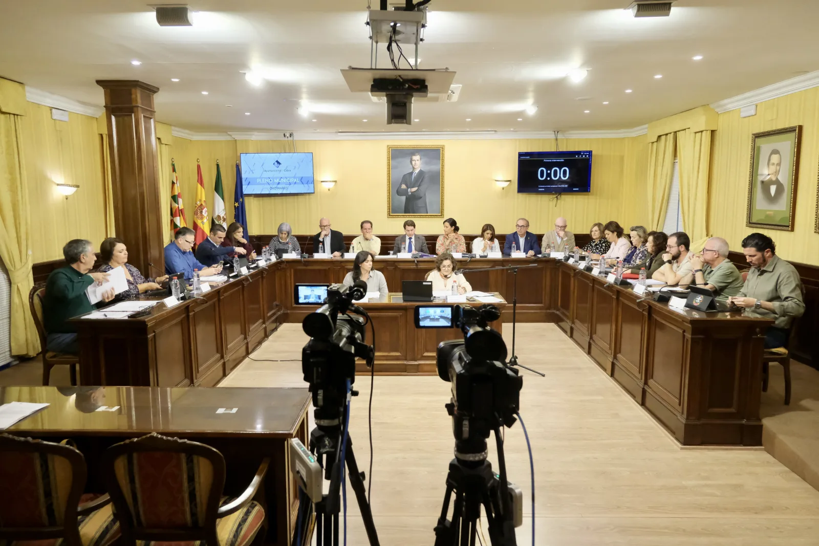 Sala de reuniones con una mesa en U y muchas personas sentadas, dos cámaras en primer plano y pantallas con un reloj a cero.
