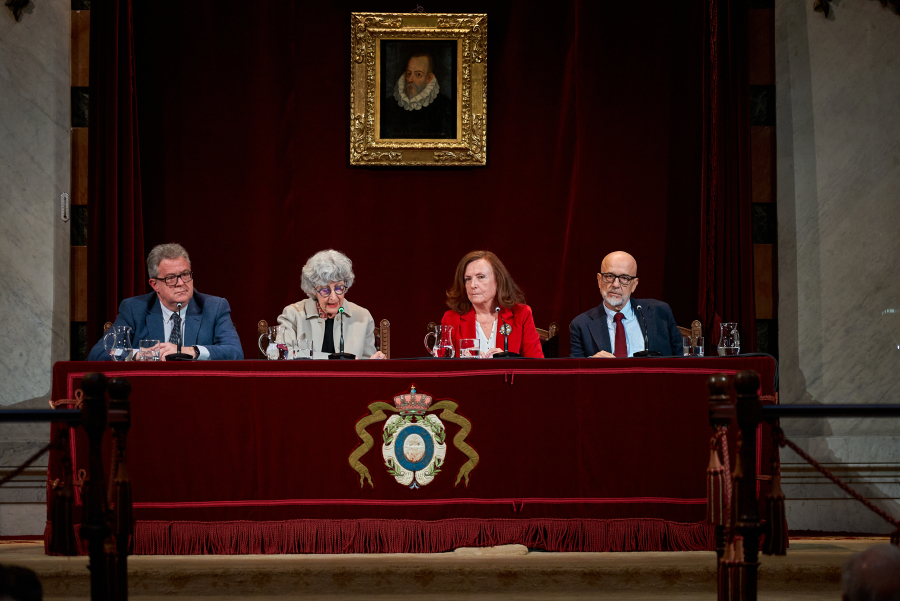 Cuatro personas sentadas tras una mesa con paño rojo y escudo, ante un retrato con marco dorado sobre cortinaje rojo.