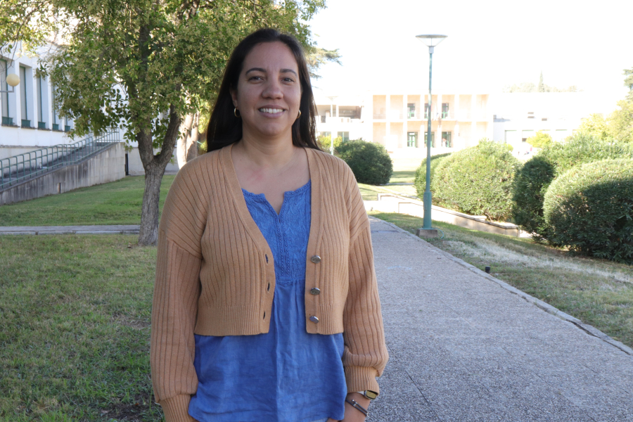 Una mujer sonriente con rebeca beige y blusa azul posa en un camino de un parque junto a árboles, setos y edificios al fondo.