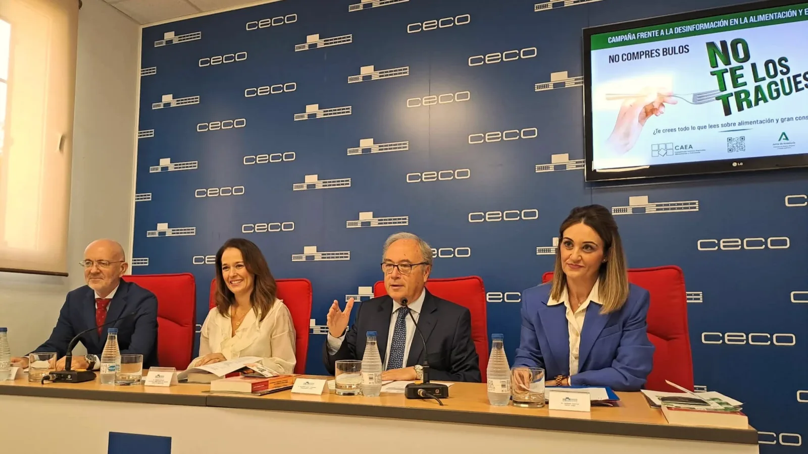 Cuatro personas sentadas tras una mesa en una sala con panel azul de logos y una pantalla con una campaña sobre alimentación.