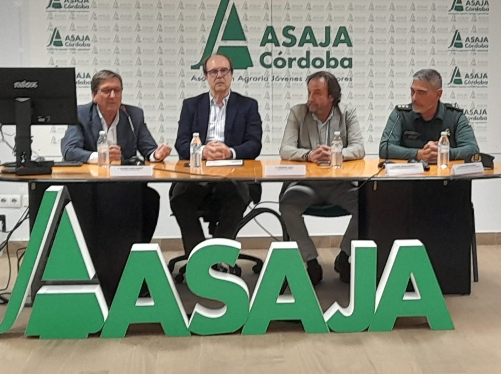 Cuatro hombres sentados tras una mesa en una sala, con un cartel grande de ASAJA en el suelo y el logotipo de ASAJA Córdoba en la pared.