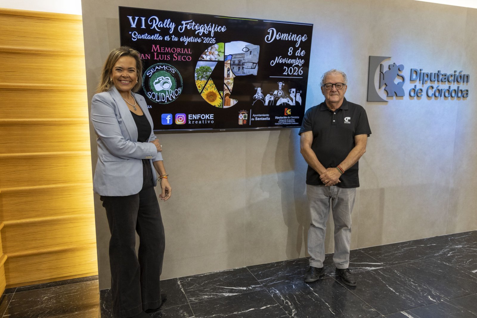 Un hombre y una mujer posan junto a una pantalla que anuncia un rally fotográfico, en una sala con el logotipo de la Diputación de Córdoba.