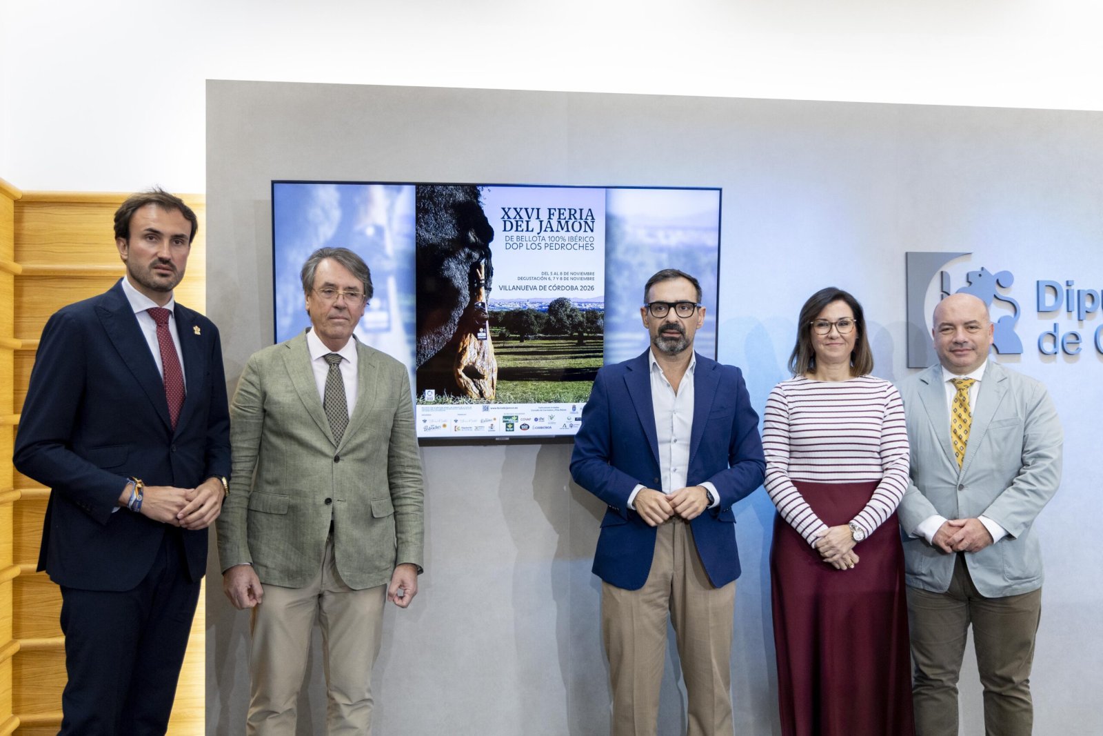 Cinco personas trajeadas posan ante una pantalla que muestra el cartel de la XXVI Feria del Jamón de Villanueva de Córdoba.