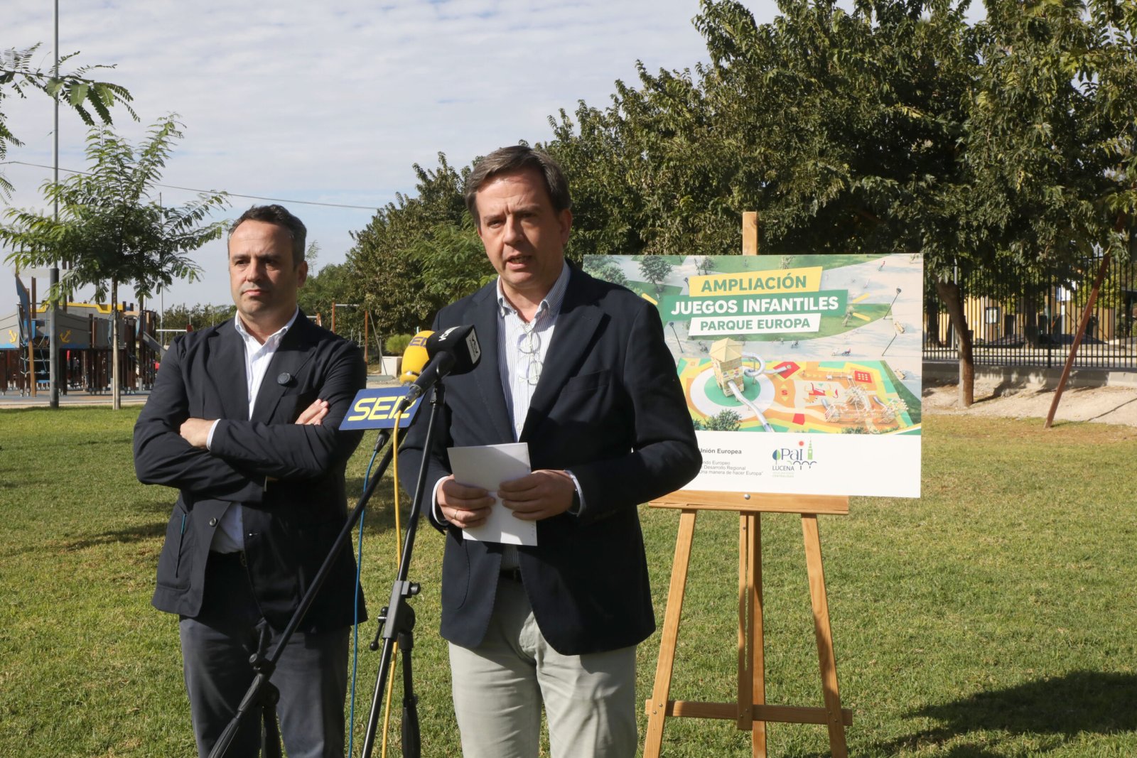 Dos hombres trajeados junto a un atril con micrófonos y un cartel sobre la ampliación de juegos infantiles en un parque.