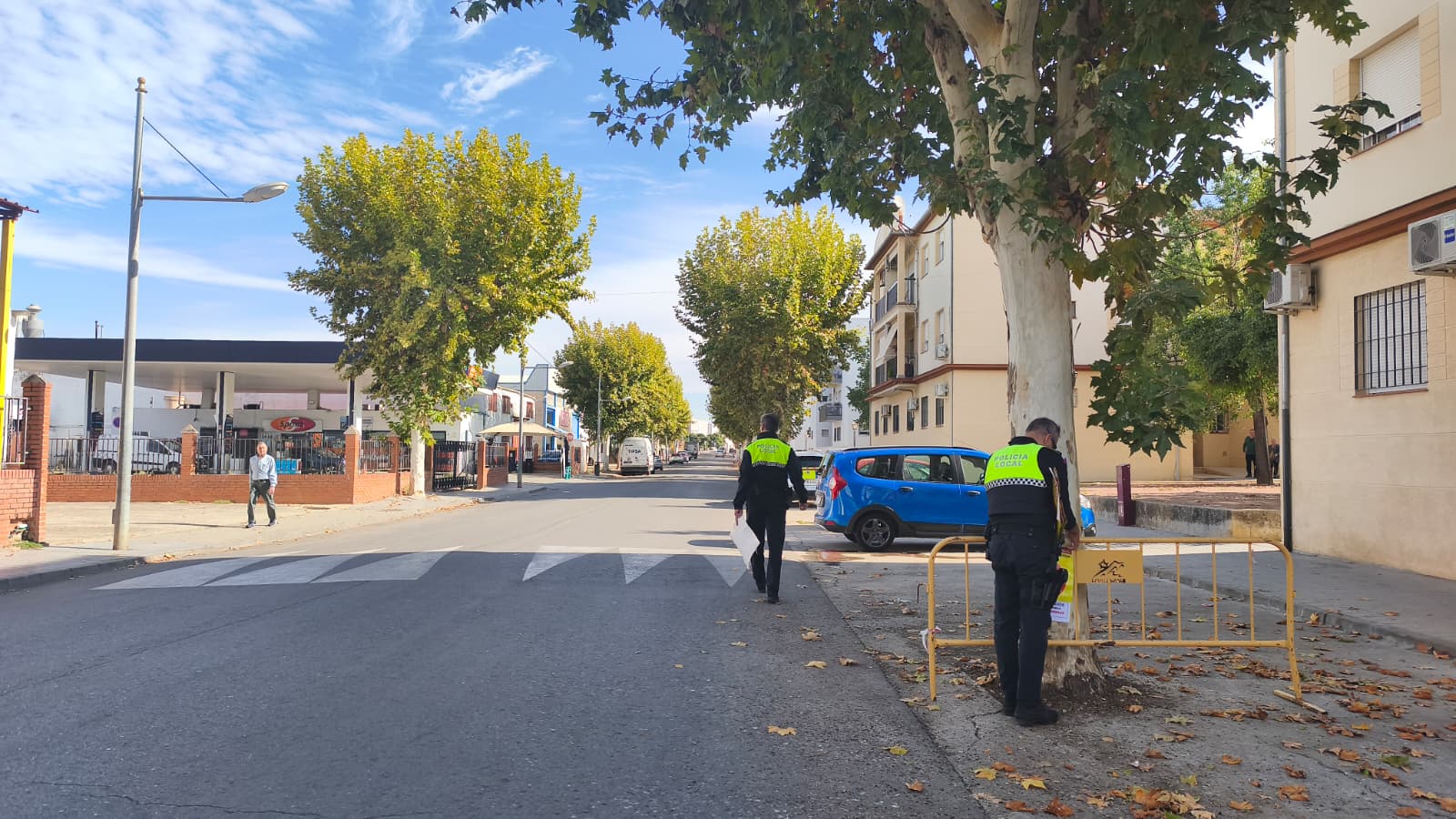 Una calle con dos agentes de policía local junto a una valla amarilla, un coche azul aparcado y hojas secas en el suelo.