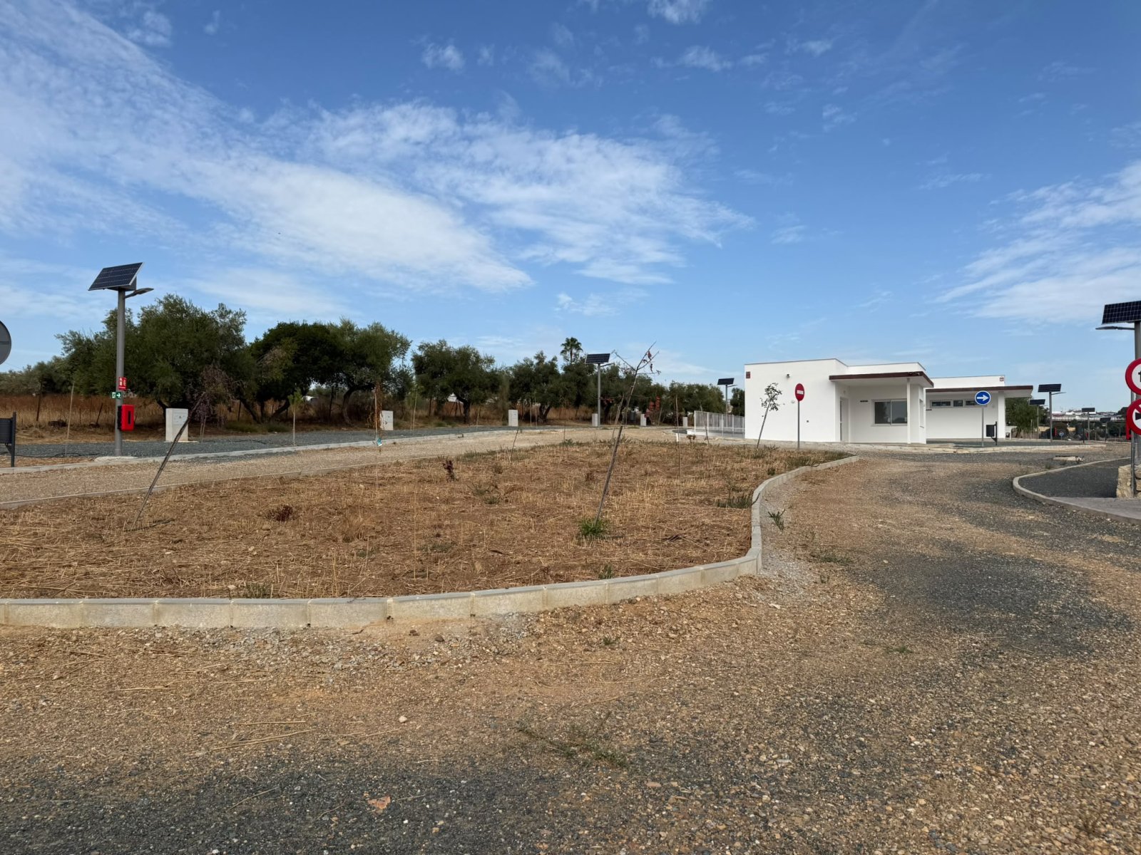 Zona de aparcamiento de grava con un parterre central seco, farolas solares y un edificio blanco bajo al fondo.