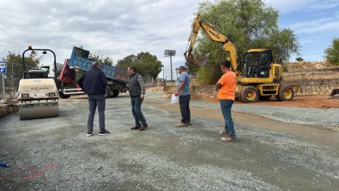 Cuatro hombres de pie sobre grava junto a una excavadora amarilla, un camión volquete y un rodillo compactador en una obra.