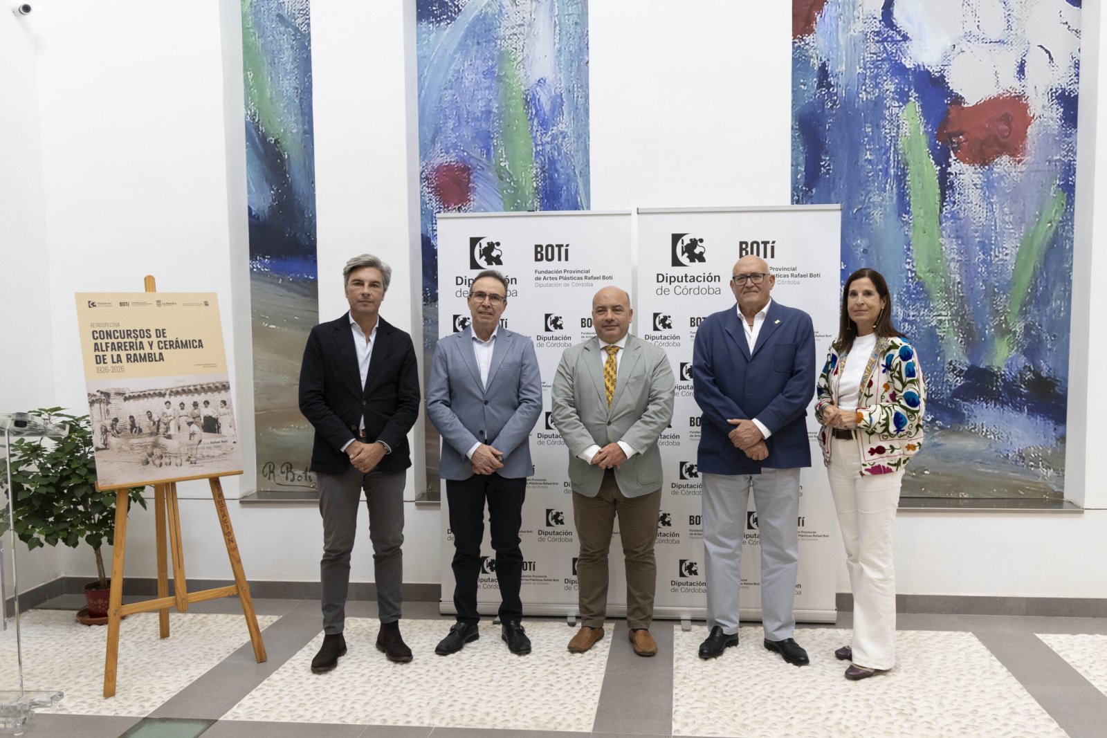Cinco personas trajeadas posan ante carteles de la Diputación de Córdoba y un cuadro abstracto azul, junto a un cartel sobre cerámica de La Rambla.