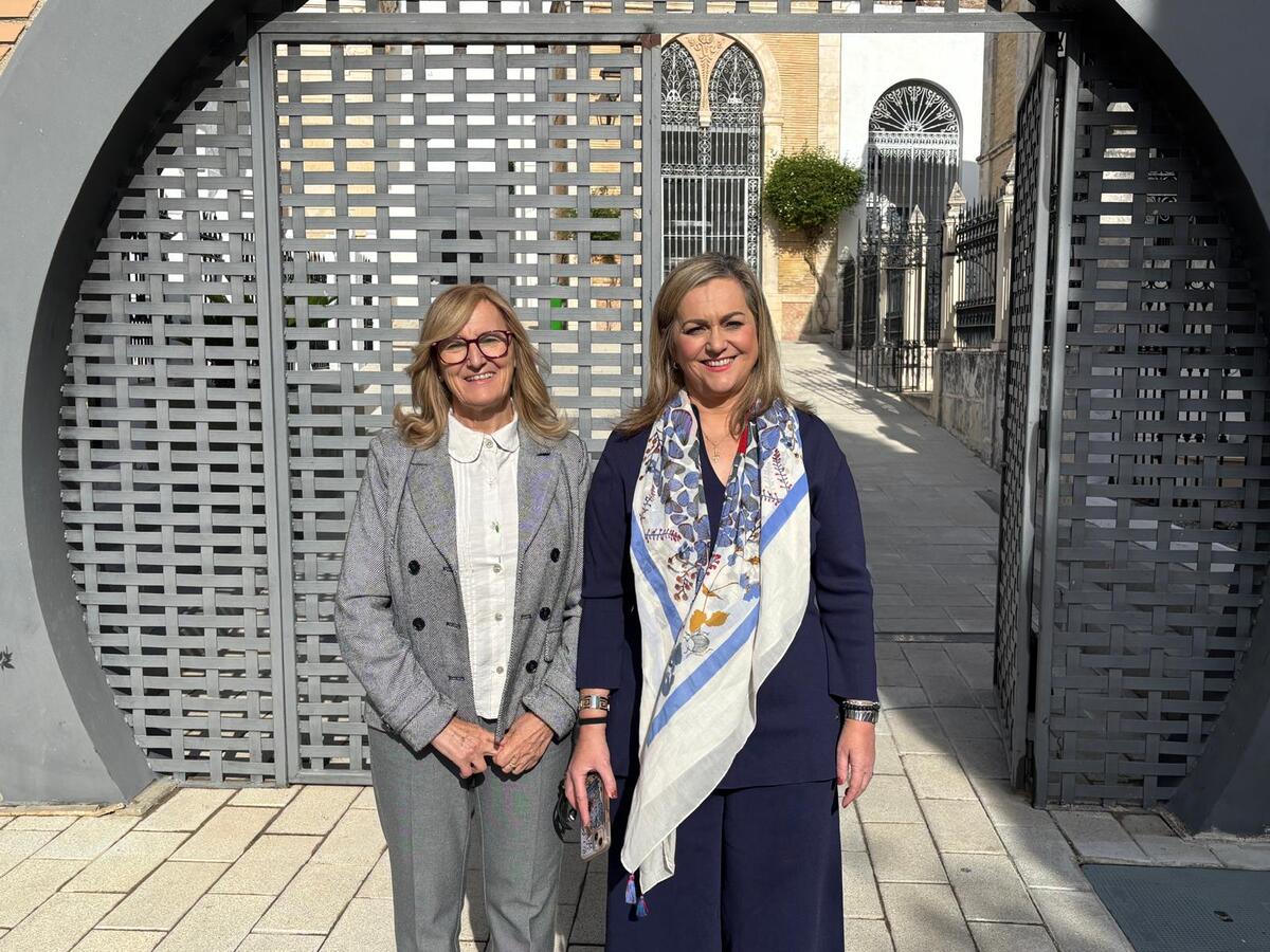 Dos mujeres sonríen de pie ante una gran puerta metálica calada, con verjas y edificios de piedra al fondo.