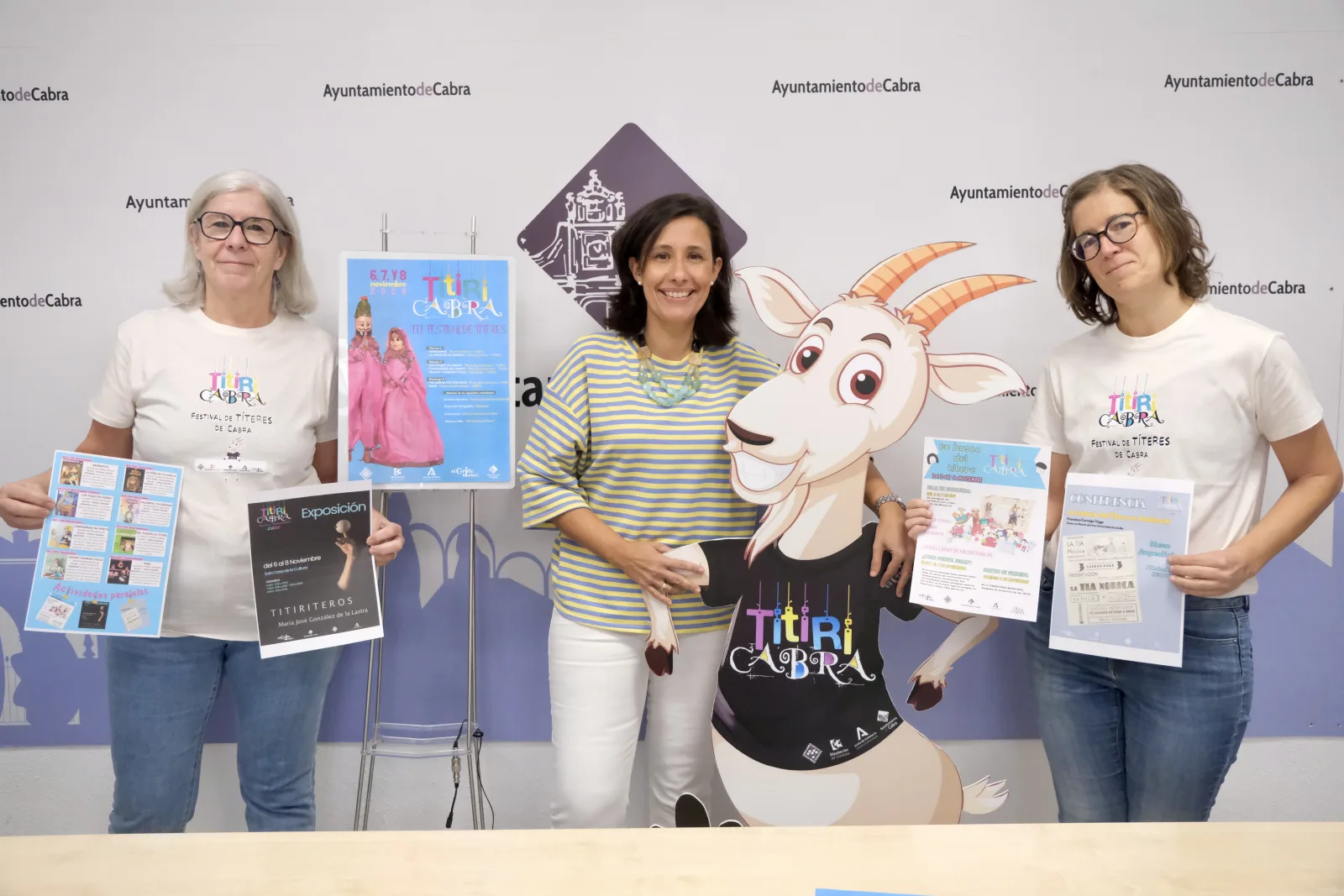 Tres mujeres posan con carteles y folletos ante un fondo con el logotipo del Ayuntamiento de Cabra y una figura recortada de una cabra.