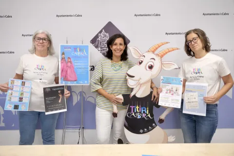 Tres mujeres de pie sostienen carteles y folletos junto a una figura recortada de una cabra, ante un fondo con el texto Ayuntamiento de Cabra.