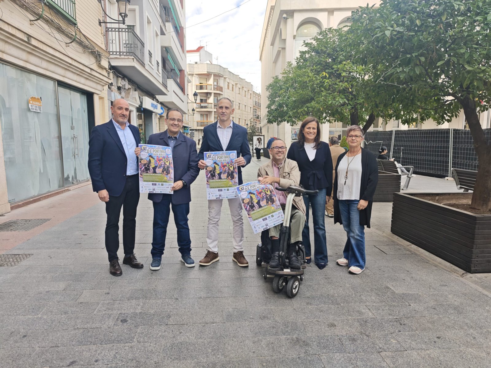 Seis personas posan en una calle peatonal; tres sostienen carteles y una está sentada en un scooter de movilidad.