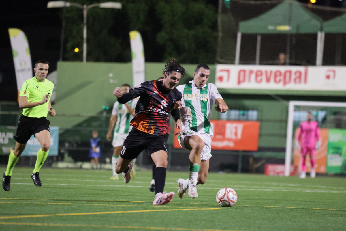 Dos futbolistas disputan el balón en un campo de hierba artificial de noche, con un árbitro y un portero al fondo.