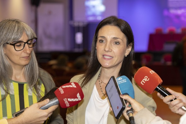 Dos mujeres en un acto; una habla rodeada de micrófonos y la otra escucha a su lado.