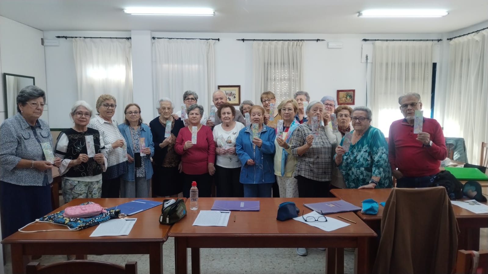 Un grupo de personas mayores posa de pie tras unas mesas en una sala, sosteniendo cada una un folleto.