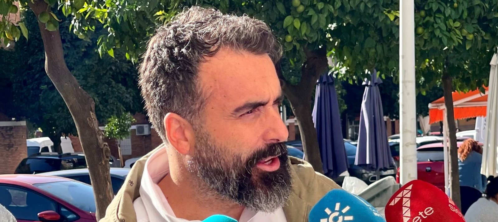 Primer plano de un hombre barbudo hablando ante varios micrófonos de medios, al aire libre junto a árboles y coches.