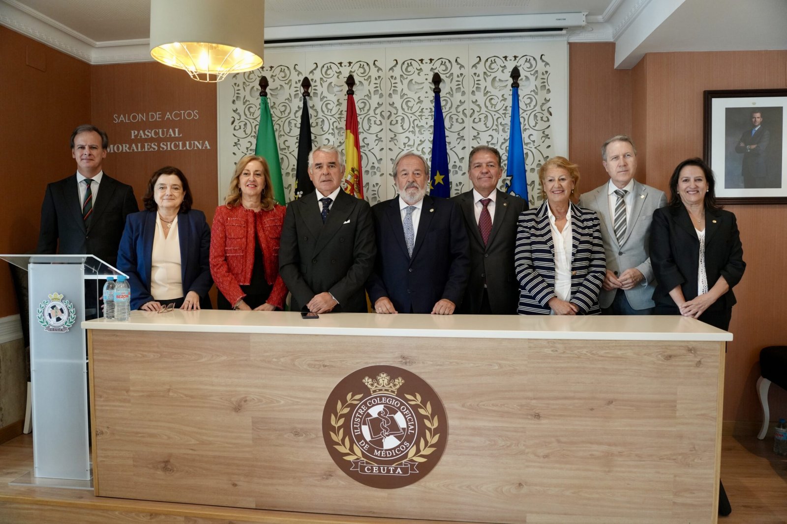 Ocho personas trajeadas posan tras una mesa con el emblema del Colegio de Médicos de Ceuta, ante banderas y un atril.