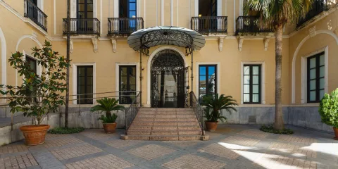 Fachada amarilla de un edificio con balcones de hierro, una escalinata central y una marquesina sobre la puerta de entrada.