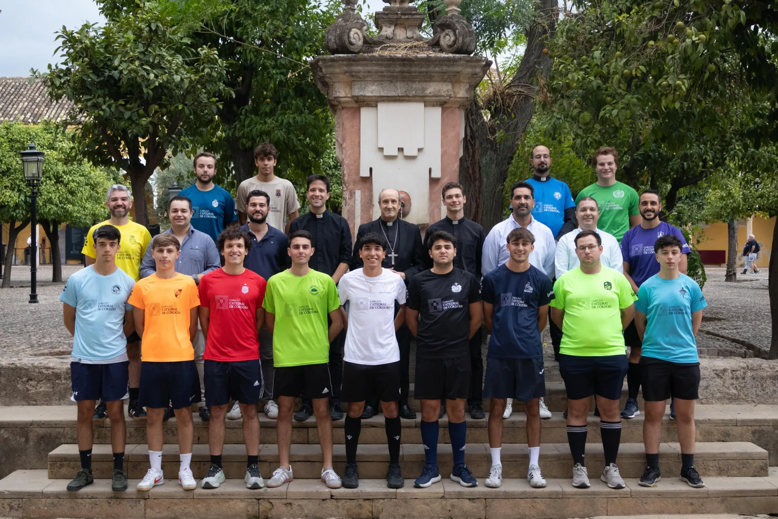 Un grupo de unas veinte personas, muchas con camisetas de colores, posa en unas escaleras junto a un monumento de piedra entre árboles.
