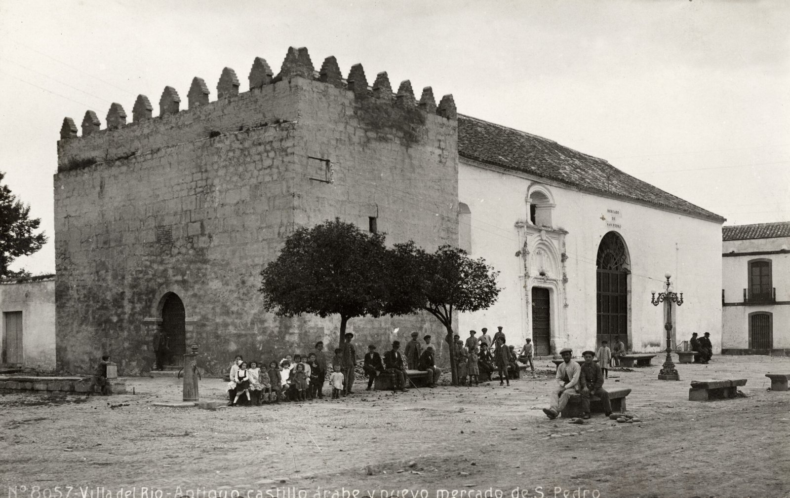 Plaza de tierra con un edificio de piedra almenado y otro blanco con arco, y un grupo de vecinos sentados o de pie junto a unos árboles.