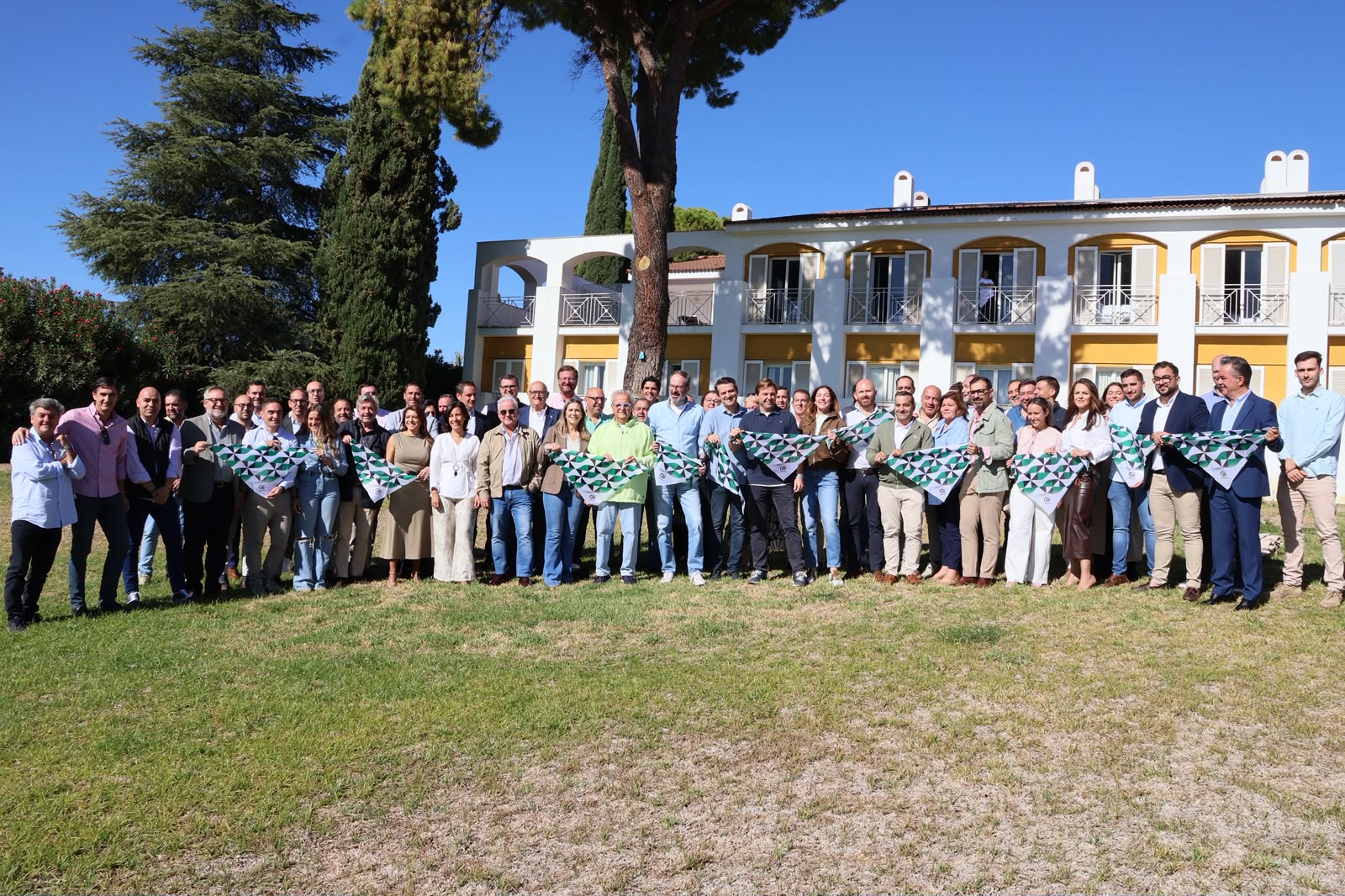 Un grupo numeroso de personas posa en un prado frente a un edificio blanco y amarillo con arcos, sosteniendo pañuelos verdes y blancos.
