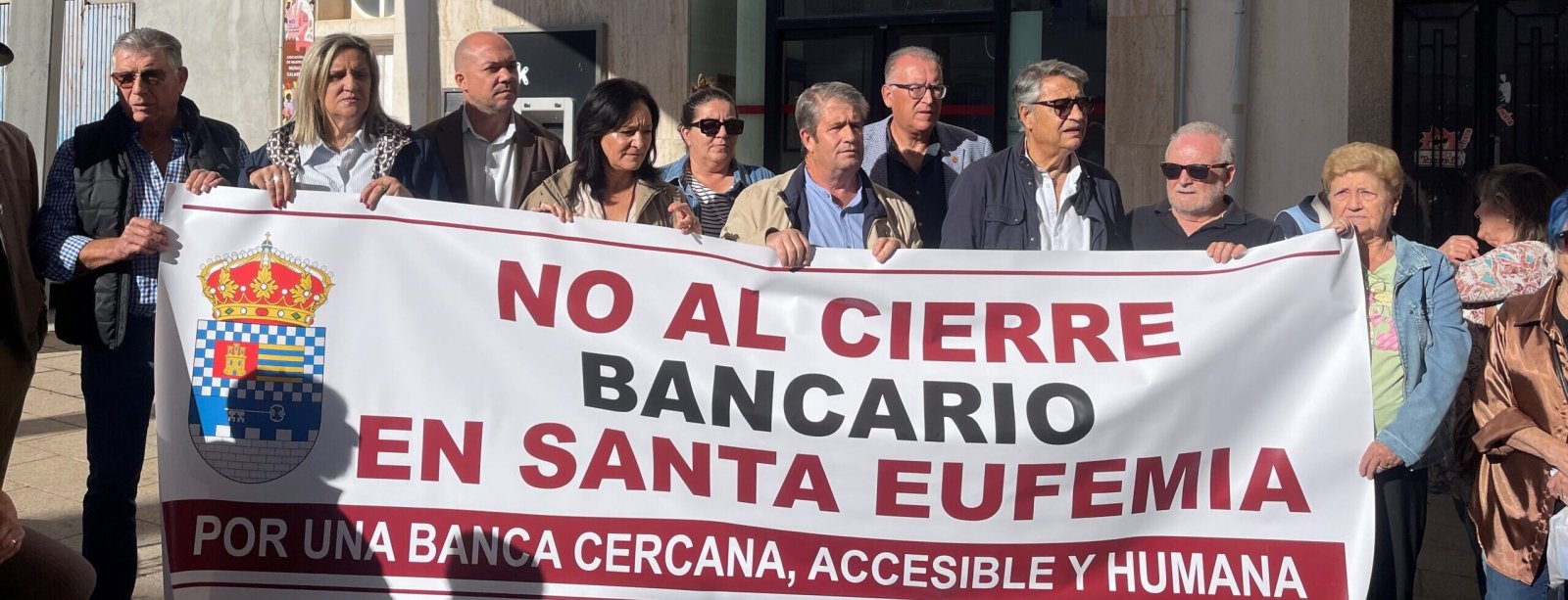 Un grupo de personas sostiene una pancarta grande que dice NO AL CIERRE BANCARIO EN SANTA EUFEMIA frente a un edificio.