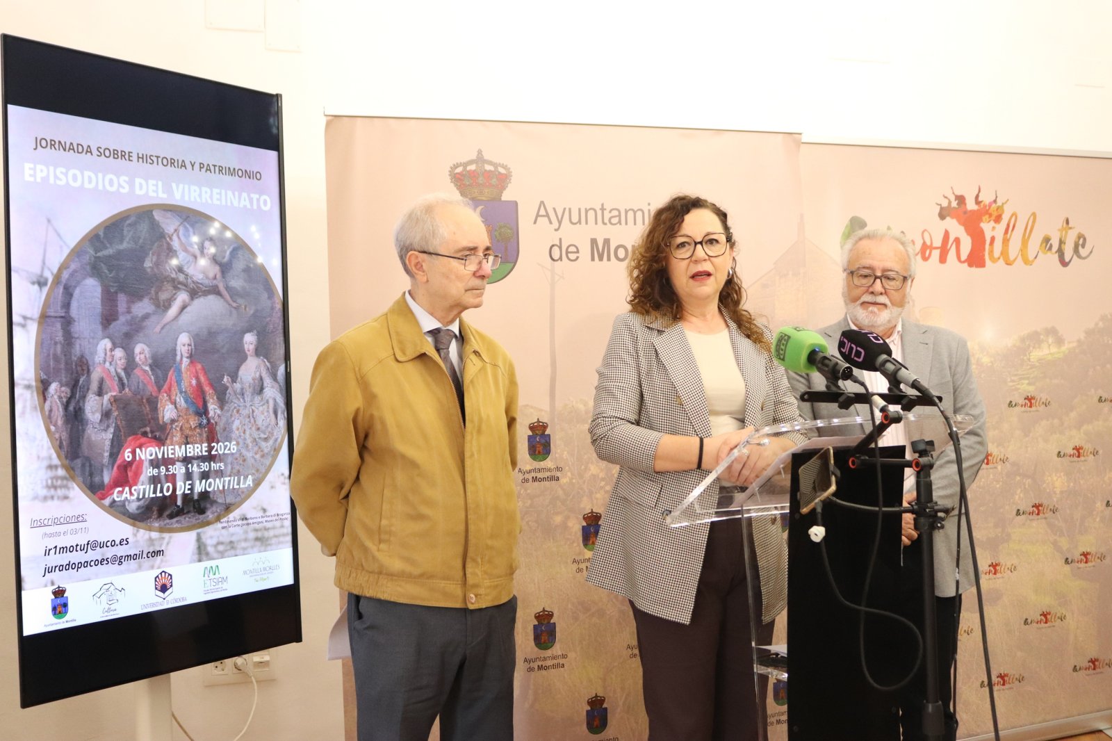 Tres personas junto a una pantalla con un cartel de jornadas históricas y un atril con micrófonos ante un fondo institucional.