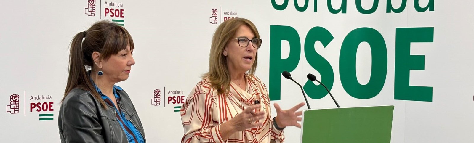 Dos mujeres ante un atril con micrófonos; una habla con las manos y la otra escucha, sobre un fondo con el logotipo PSOE Andalucía.
