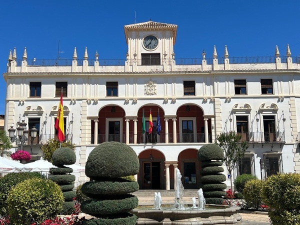 Edificio municipal de fachada blanca con reloj central, arcos y banderas, y una fuente con setos recortados delante.