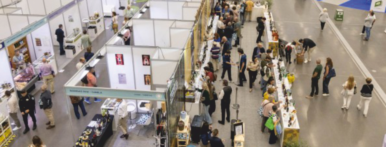Vista elevada de una feria con puestos blancos y pasillos donde muchas personas recorren y atienden expositores de productos.