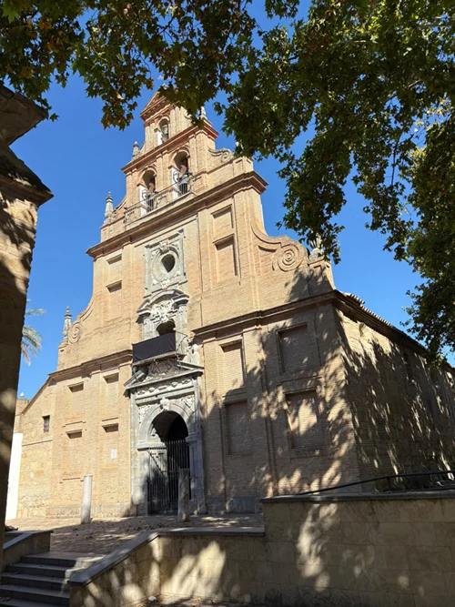 Fachada de piedra de una iglesia con portada barroca, hornacinas y una torre escalonada, vista entre árboles bajo cielo azul.
