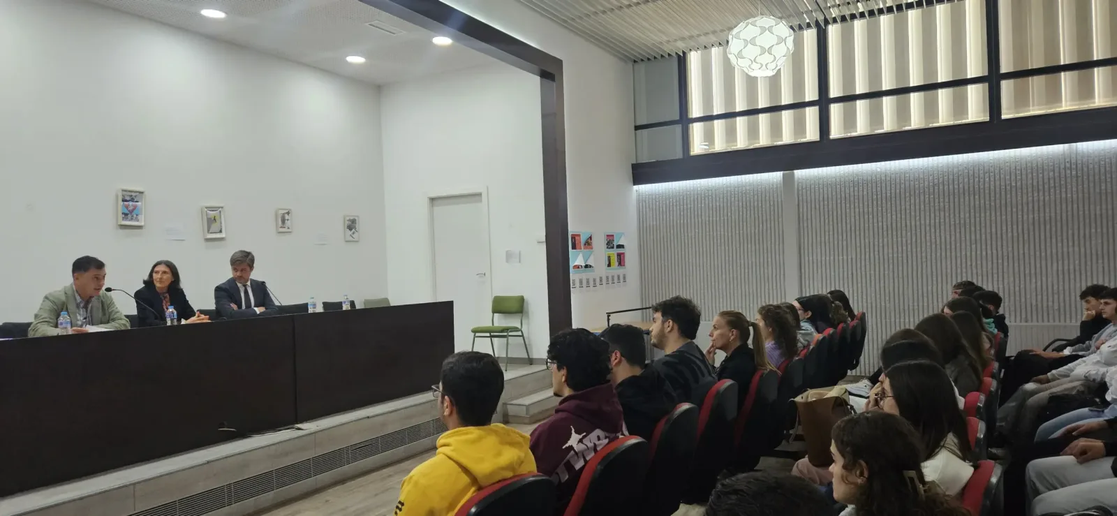 Tres personas sentadas tras una mesa presiden ante un público joven sentado en un aula.