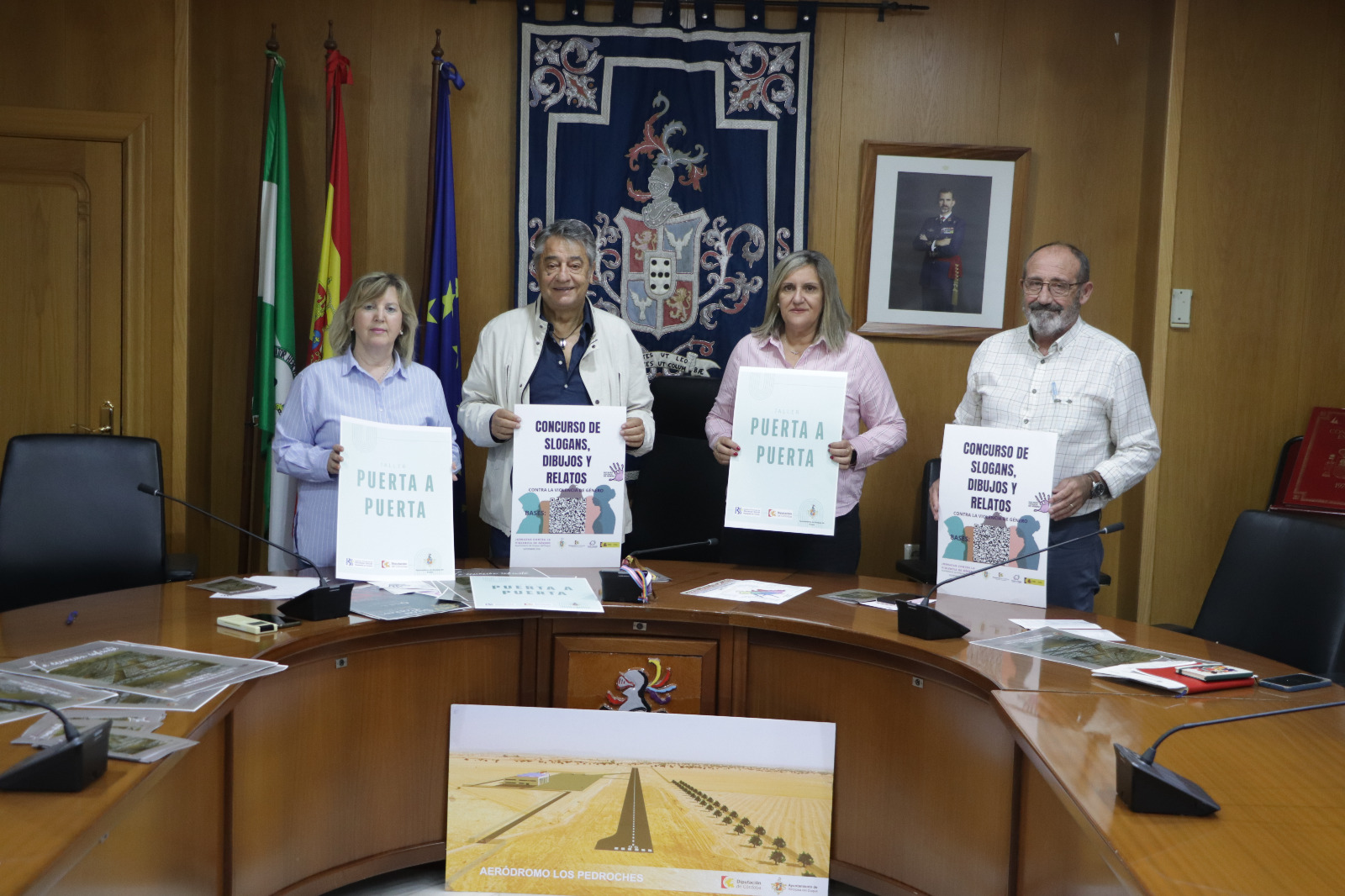 Cuatro personas de pie tras una mesa ovalada sostienen carteles con lemas de un concurso, en una sala con banderas y un escudo.