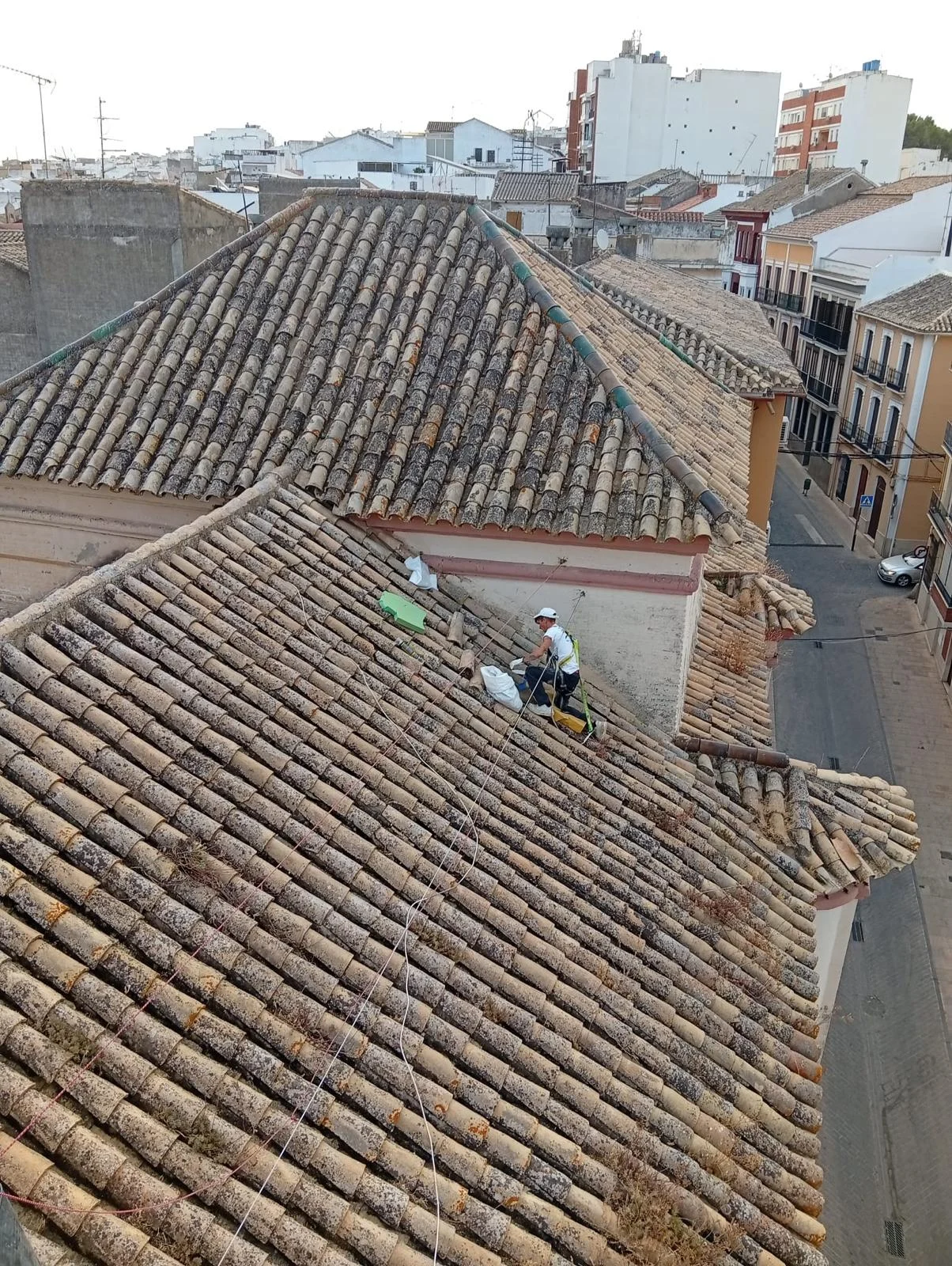 Un operario con casco y arnés trabaja sobre un tejado de tejas junto a sacos, con edificios y una calle al fondo.