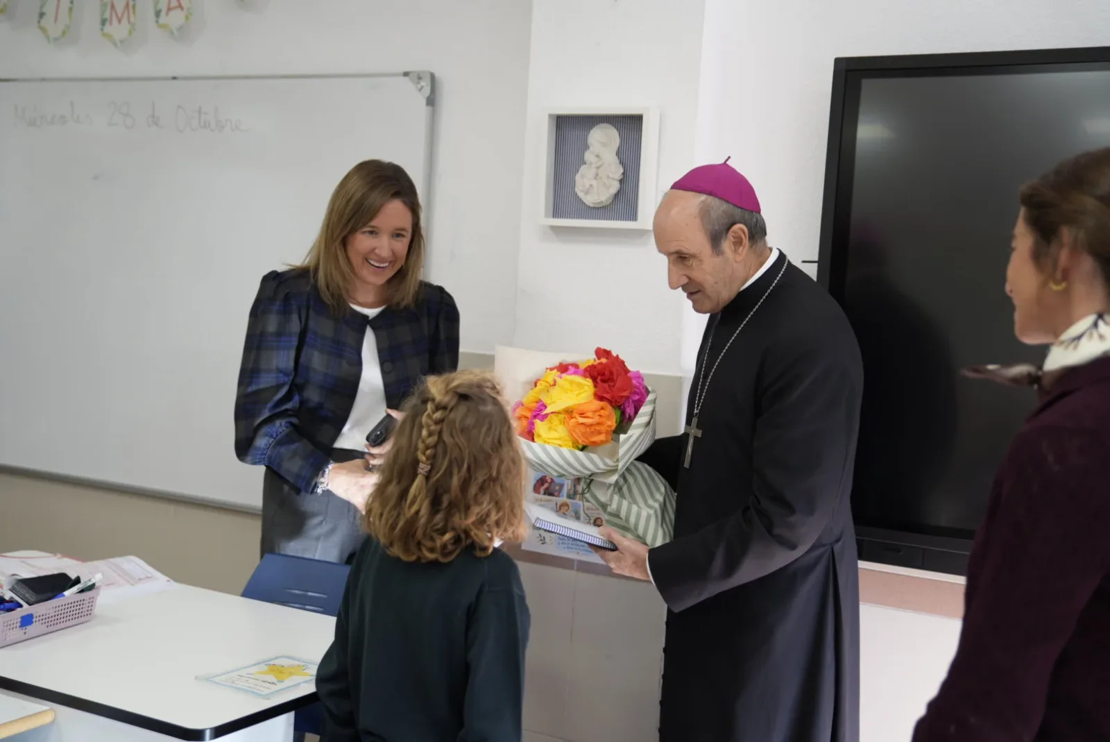 Un hombre con solideo morado y cruz recibe un ramo de flores de una niña en un aula, ante dos mujeres sonrientes.