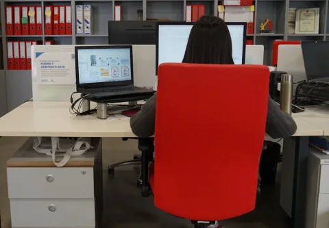 Una persona de espaldas, con melena oscura, sentada en una silla roja frente a un escritorio con un portátil y una pantalla, en una oficina con estanterías de a