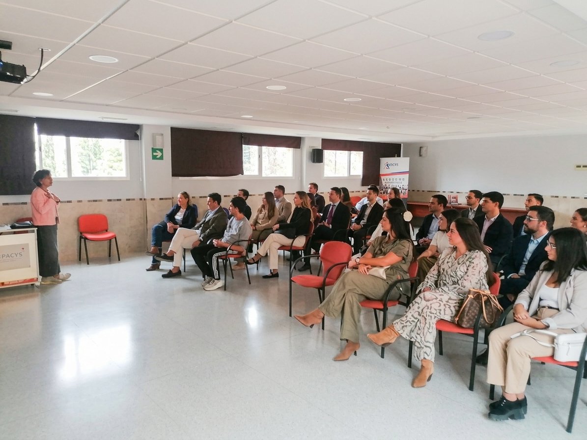 Una mujer habla ante un grupo de personas sentadas en sillas rojas en una sala amplia con ventanas.