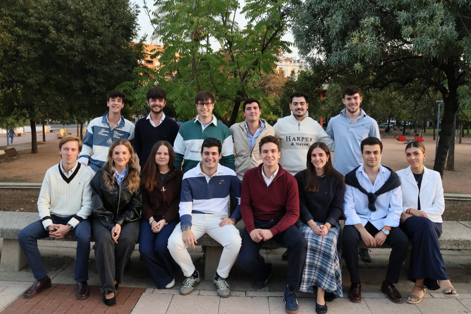 Un grupo de trece jóvenes, seis de pie y siete sentados en un banco de piedra, posan juntos en un paseo arbolado.